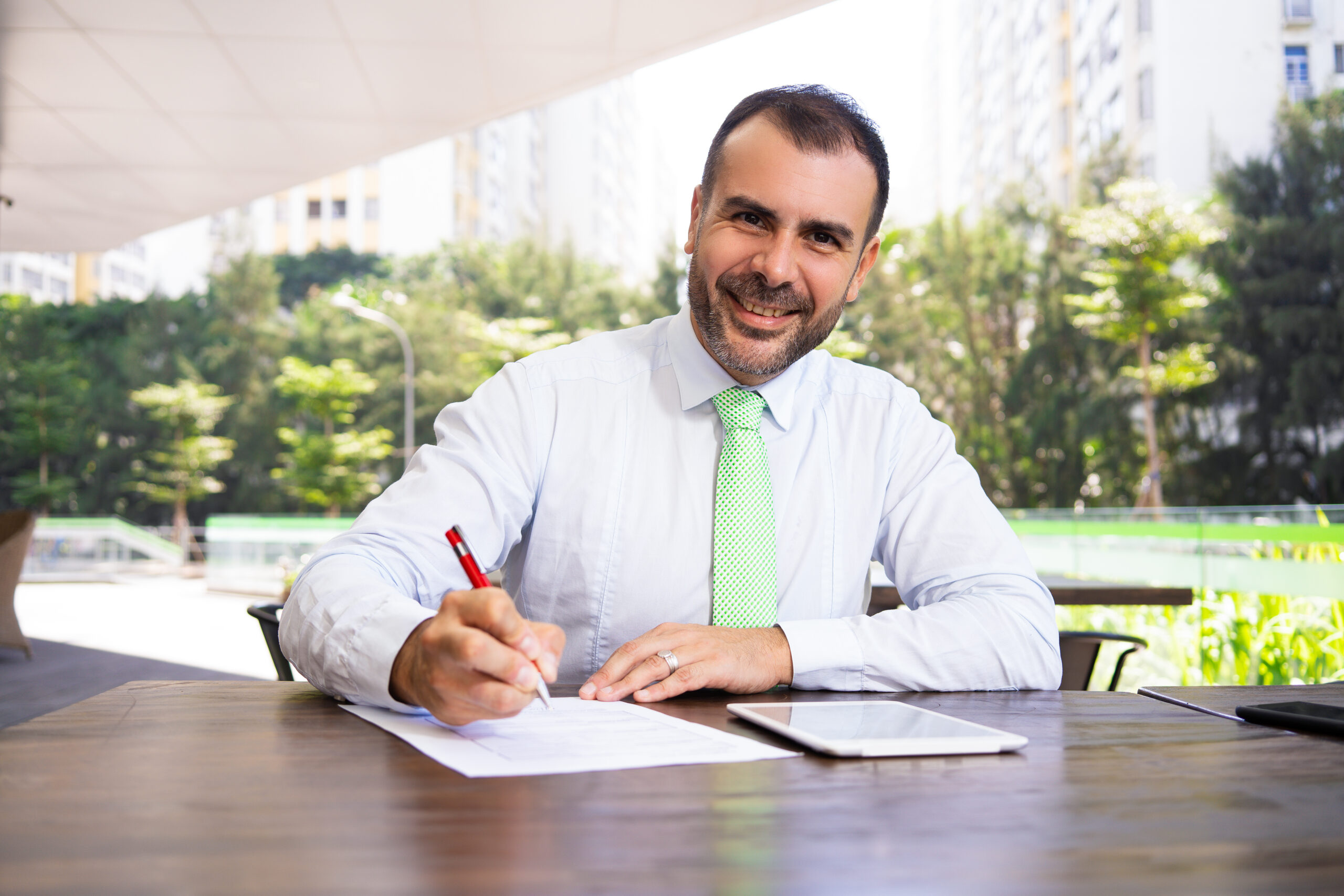 Portrait of smiling mature businessman signing agreement outdoors. Caucasian bearded entrepreneur sitting at cafe table, looking at camera and smiling. Successful businessman concept